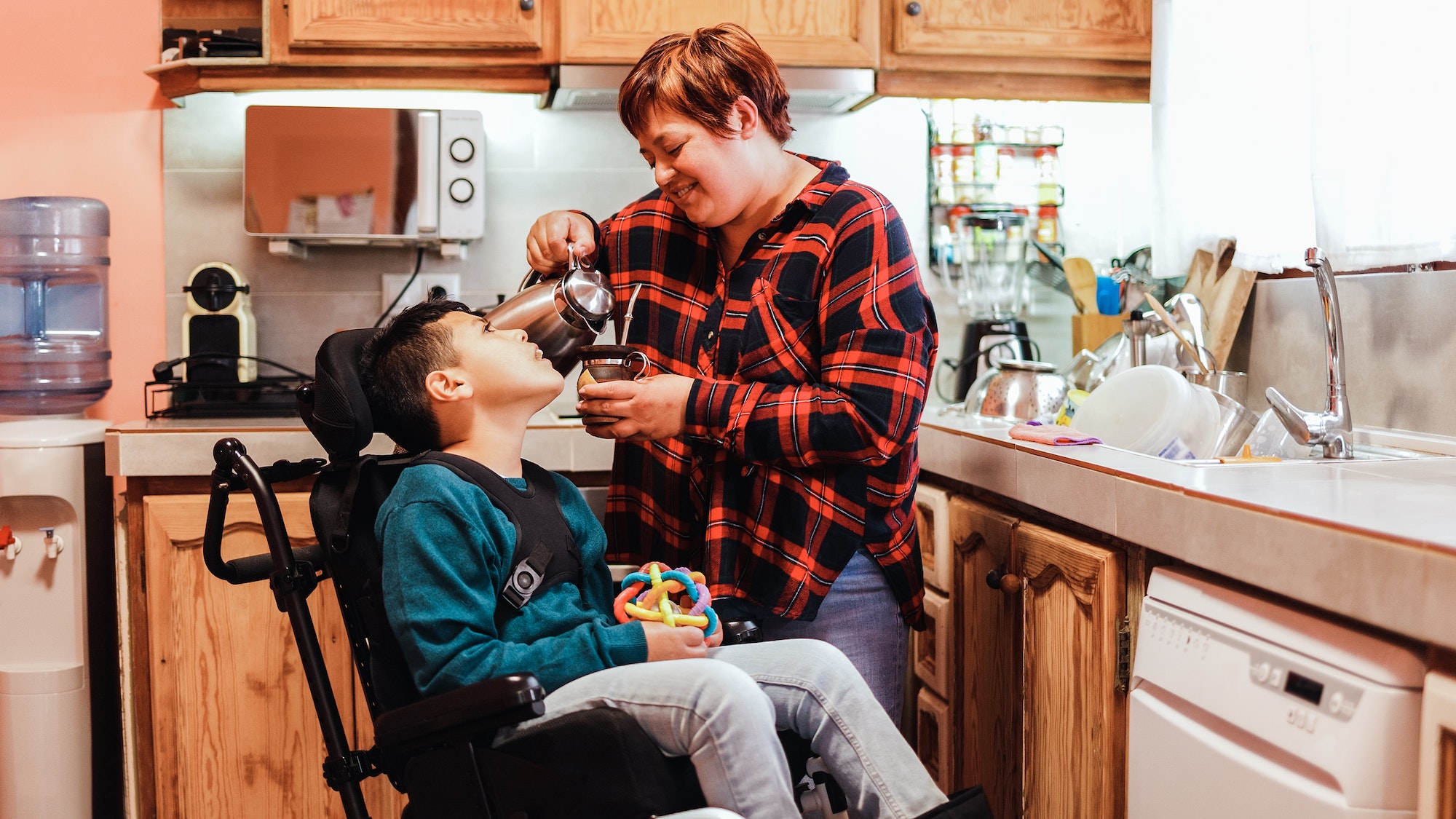 Latin mother taking care of son with disability on wheelchair at home kitchen - Focus on mom face